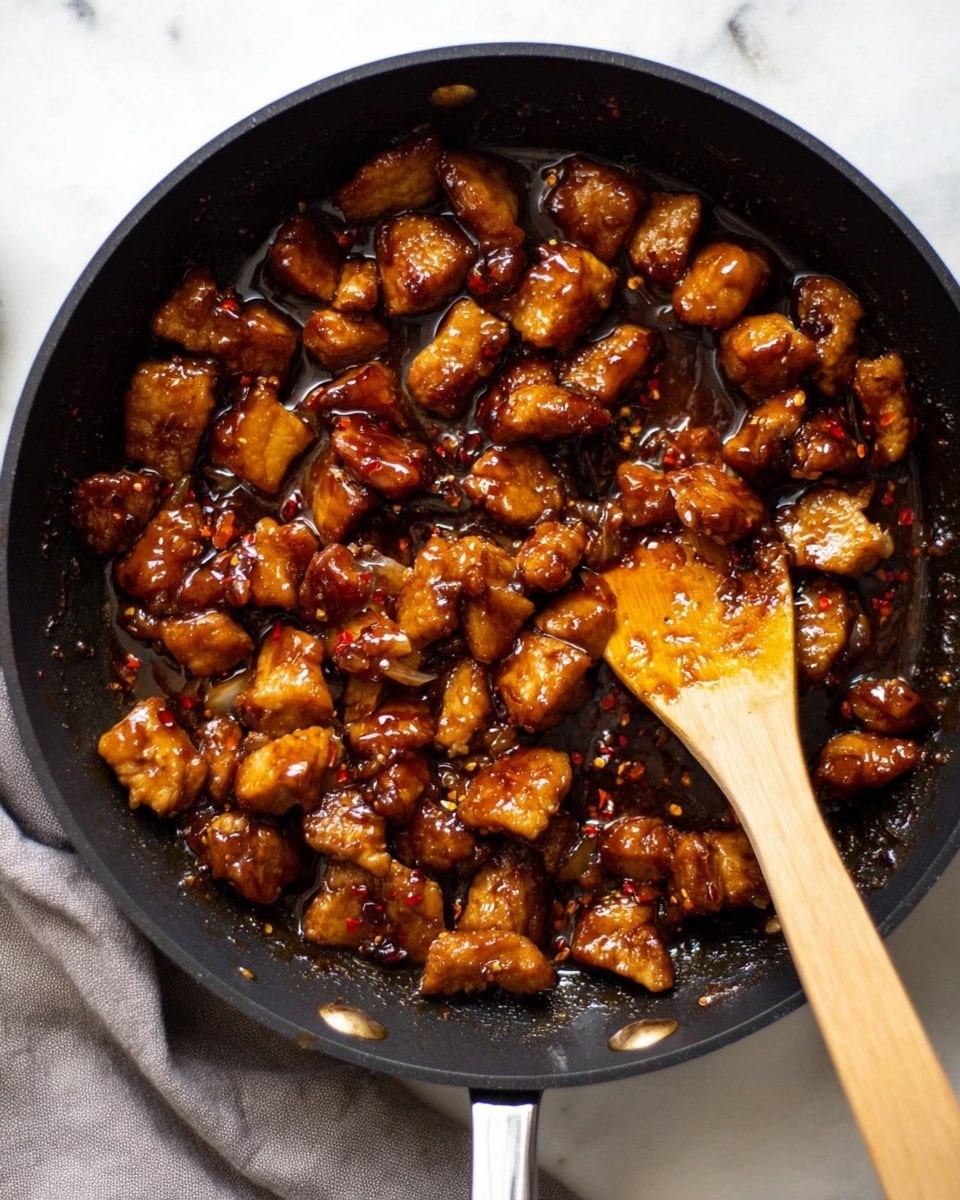 The image shows a black pan filled with many small, golden brown pieces of meat covered in a shiny, thick dark brown sauce with hints of red chili flakes. The meat pieces vary in size and texture, some look soft while others are slightly crispy. A light brown wooden spatula is placed inside the pan, partially stirring the meat and sauce. The pan rests on a white marbled surface with a corner of a gray cloth visible below it. The light in the photo highlights the glaze on the meat, making it look shiny and delicious. photo taken with an iphone --ar 4:5 --v 7