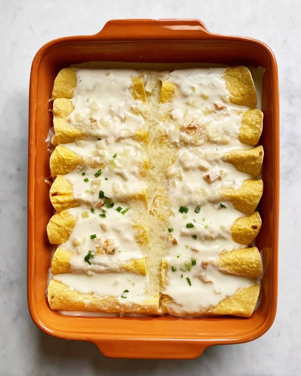 A rectangular orange baking dish holds two rows of rolled yellow tortillas filled with a light-colored filling, topped with a smooth, thick white sauce that slightly covers the edges of the tortillas and has small bits of green chili scattered throughout; the dish rests on a white marbled surface. photo taken with an iphone --ar 4:5 --v 7