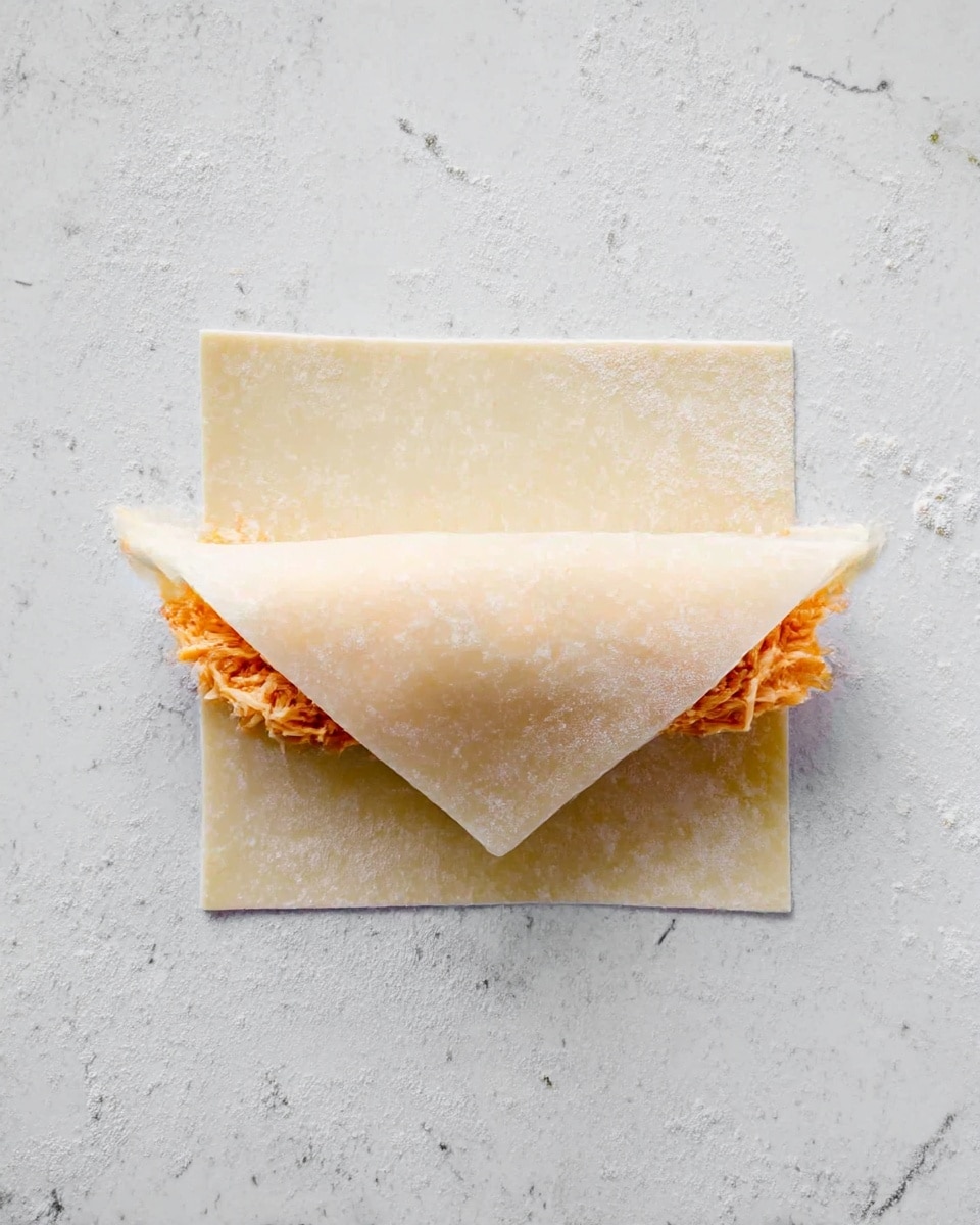 A single thin, square, pale dough wrapper is placed flat on a white marbled surface, folded slightly upwards from the bottom edge to cover part of the filling. The filling inside is orange and shredded, peeking out on both left and right sides just above the folded edge of the dough, showing a moist and soft texture. The dough is smooth and lightly textured with a translucent quality, allowing a faint pattern from the surface beneath to show through. Photo taken with an iphone --ar 4:5 --v 7