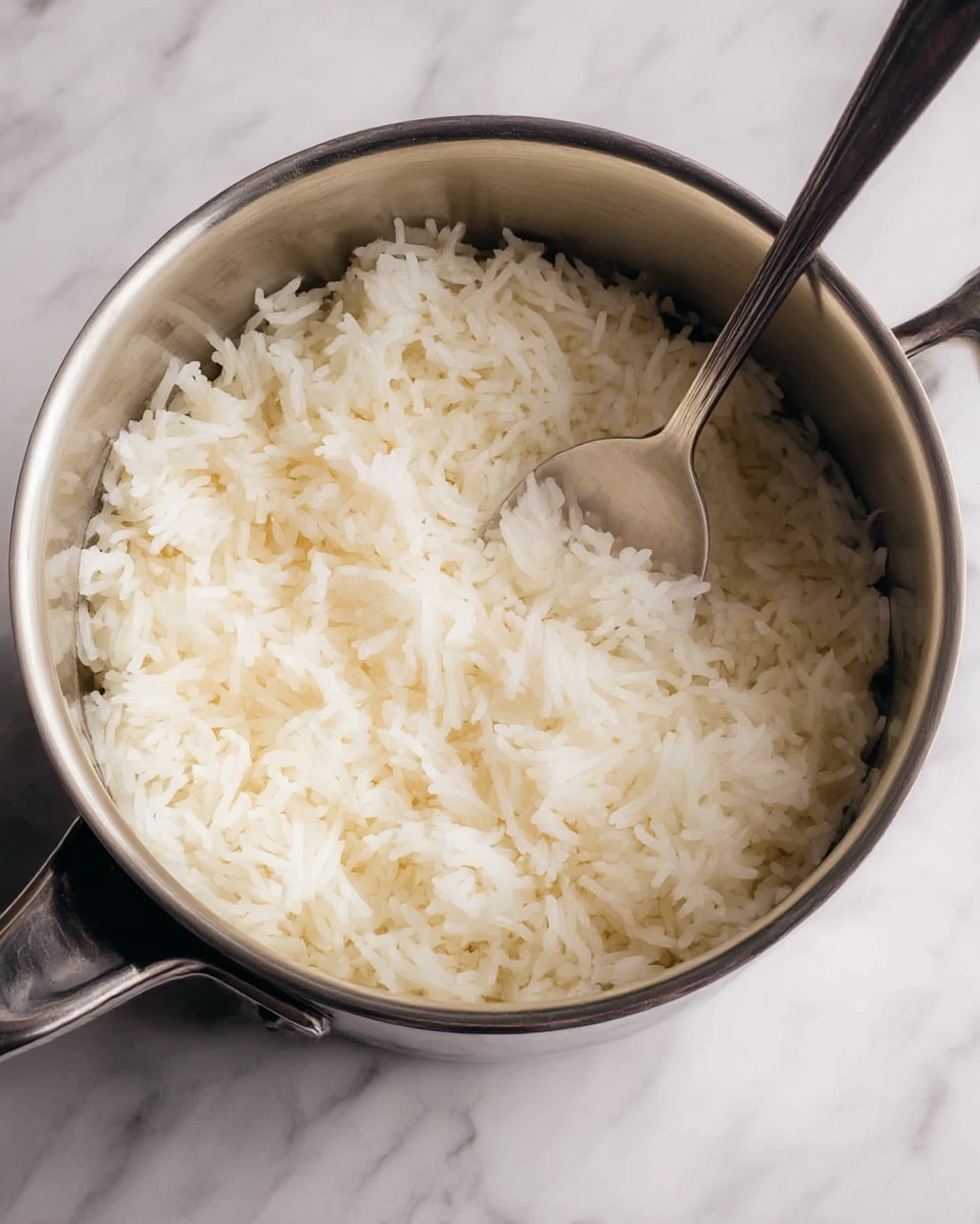 A close-up image of a stainless steel pot filled with cooked white rice, with the grains fluffy and soft, showing a light texture. Inside the pot, there is a metal spoon partially buried in the rice, its handle resting against the pot's edge. The pot has a shiny metallic surface, and the background features a white marbled texture. photo taken with an iphone --ar 4:5 --v 7