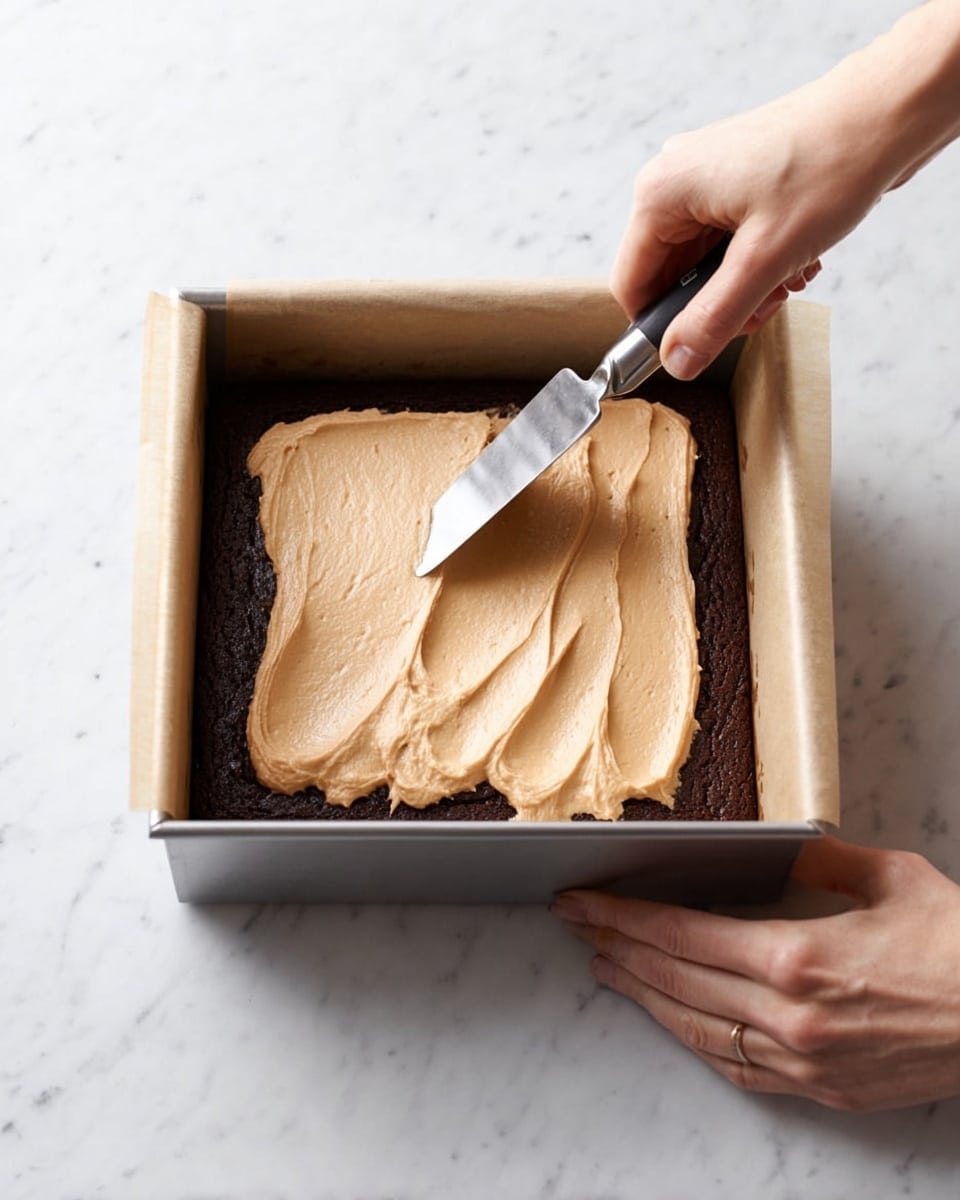 A square metal baking pan lined with parchment paper holds a dark brown baked base layer, which is dense and smooth in texture. On top, a thick layer of light tan, creamy frosting is being evenly spread with an angled metal spatula held by a woman's hand from above. Another woman's hand is steadying the pan from the right side. The scene is set on a clean white marbled surface. The light softly highlights the smooth and slightly glossy surface of the frosting against the matte texture of the baked layer. Photo taken with an iphone --ar 4:5 --v 7