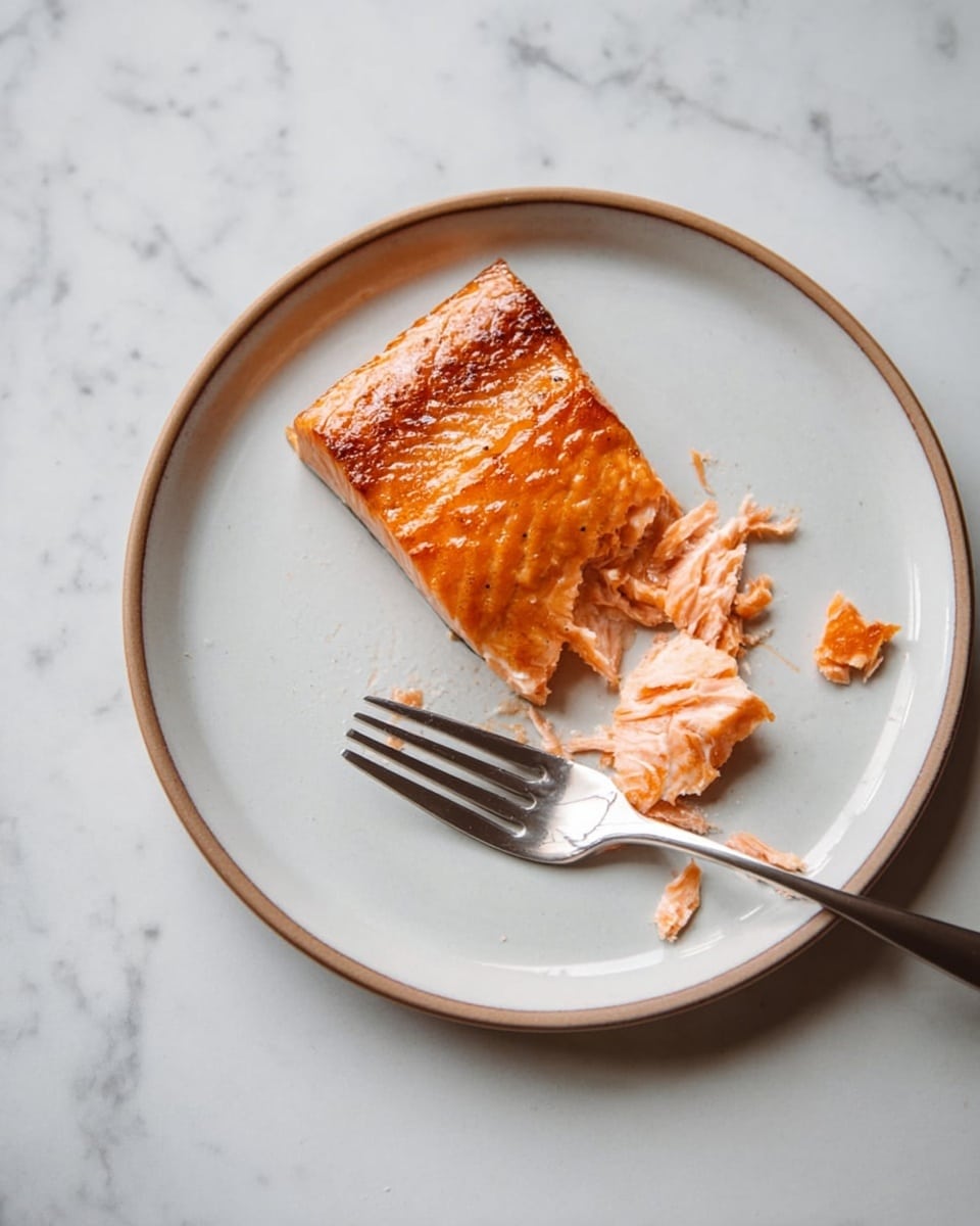 A single piece of cooked salmon with a crispy, golden-brown skin on top sits slightly off-center on a white plate with a light brown rim. The salmon's flaky, pink flesh is partially pulled apart near its lower right side, with small pieces scattered around. A silver fork rests on the plate, its tines touching some flakes of salmon. The plate is placed on a white marbled surface, showing soft gray veins. The lighting is soft and natural, highlighting the texture and color contrast between the salmon skin and flesh. photo taken with an iphone --ar 4:5 --v 7