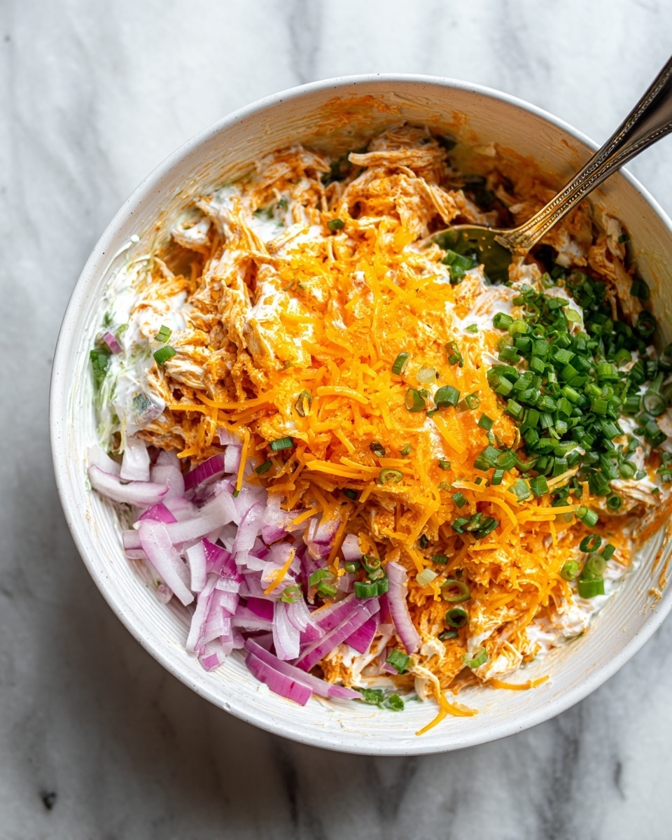 A deep white bowl holds a mixed dish with visible layers starting with shredded orange-colored chicken mixed with creamy white dressing, topped with bright orange shredded cheddar cheese. On one side, there are fresh sliced red onions with white and purple colors, while chopped green onions add a vivid green touch near the center. The dish appears slightly messy with the dressing partly stirred, showing creamy white texture combined with the orange chicken. A metal spoon is partially inside the bowl, mixing the contents. The bowl rests on a white marbled surface. Photo taken with an iphone --ar 4:5 --v 7