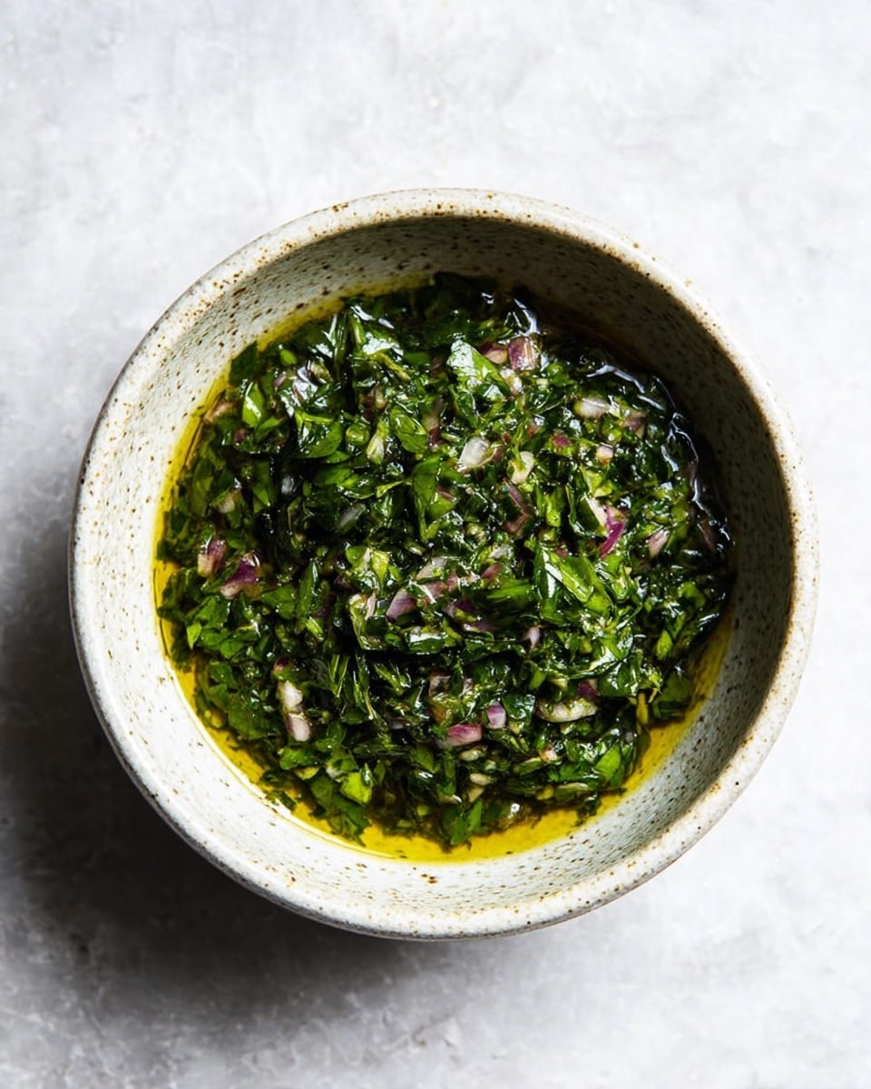 A white speckled bowl filled with a fresh green herb sauce. The sauce has finely chopped green leaves, small bits of onions with a pinkish tint, all sitting in a shiny layer of golden olive oil. The herbs and onions create a textured, slightly chunky surface with bright green and pale purple colors mixed evenly. The bowl is on a white marbled surface, showing soft natural light and gentle shadows around it. photo taken with an iphone --ar 4:5 --v 7