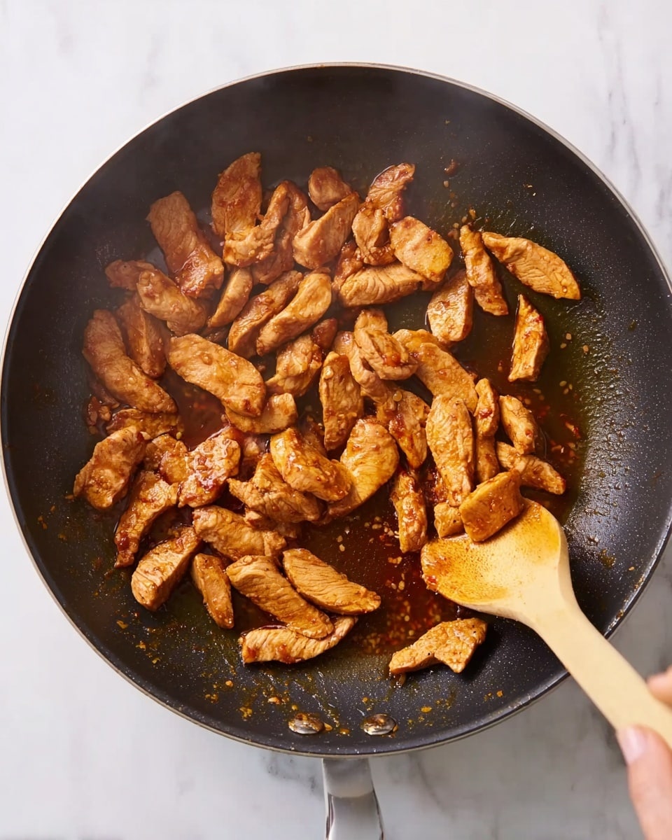A close-up view of a dark non-stick frying pan filled with small, light brown strips of cooked meat in a reddish sauce. A wooden spoon is stirring the pieces in the middle of the pan, held by a woman's hand. The pan sits on a white marbled surface, and the meat has a slightly shiny, cooked texture with some sauce pooling around the edges. steam rises slightly, showing heat and freshness. photo taken with an iphone --ar 4:5 --v 7