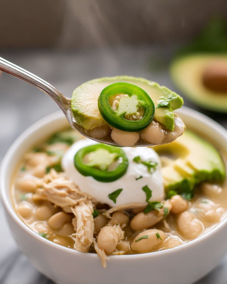 The image shows a close-up of a white bowl filled with a light broth soup containing white beans and shredded light brown chicken. On the top layer, there are several slices of green jalapeño peppers and three slices of bright green avocado, along with a small dollop of white sour cream in the center, sprinkled with small green cilantro leaves. The soup looks fresh and warm, as steam rises in the background, with a woman's hand holding a spoonful of the soup that includes beans, chicken, a jalapeño slice, sour cream, and cilantro. The bowl rests on a white marbled surface with a blurred background. photo taken with an iphone --ar 4:5 --v 7