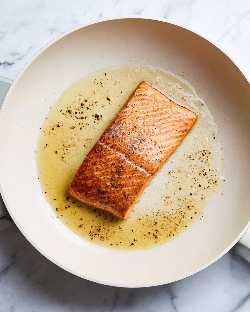 A single piece of cooked salmon with a golden brown, slightly crispy skin sits in the middle of a white frying pan. The salmon has a warm pinkish-orange color with grill marks and a texture that shows the cooked flesh layers. Around the salmon, there is a thin layer of golden oil with specks of black pepper spread across the pan. The pan is placed on a surface with a white marbled texture. photo taken with an iphone --ar 4:5 --v 7