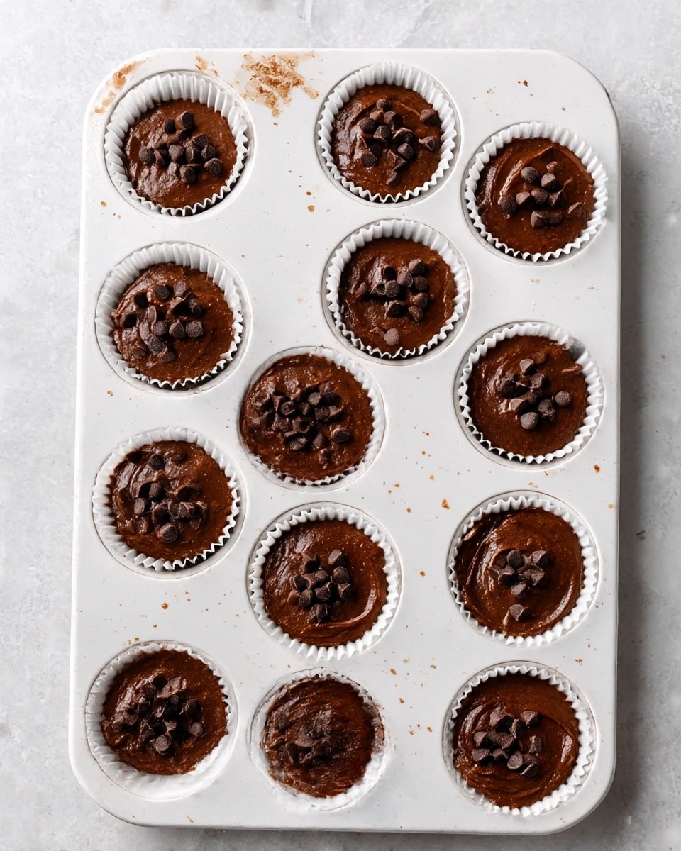 A white baking tray holds twelve cavities, each lined with white paper cups filled with smooth, dark brown chocolate batter. The top of each batter layer is sprinkled with small, dark chocolate chips scattered unevenly. The tray sits on a white marbled surface, showing some small splashes of the batter around the cavities. The texture of the batter looks creamy and thick, ready for baking. photo taken with an iphone --ar 4:5 --v 7