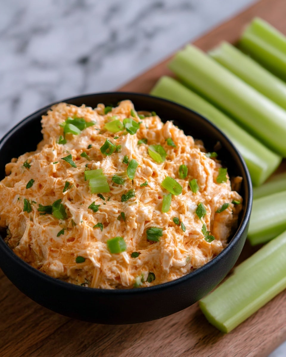 The image shows a black bowl filled with a creamy, light orange mixture with a soft, shredded texture. Small pieces of green herbs are sprinkled evenly on top, adding a fresh color contrast. The bowl sits on a wooden surface, and beside it are several pale green celery sticks arranged neatly. The background is a white marbled texture, creating a clean and simple look. photo taken with an iphone --ar 4:5 --v 7