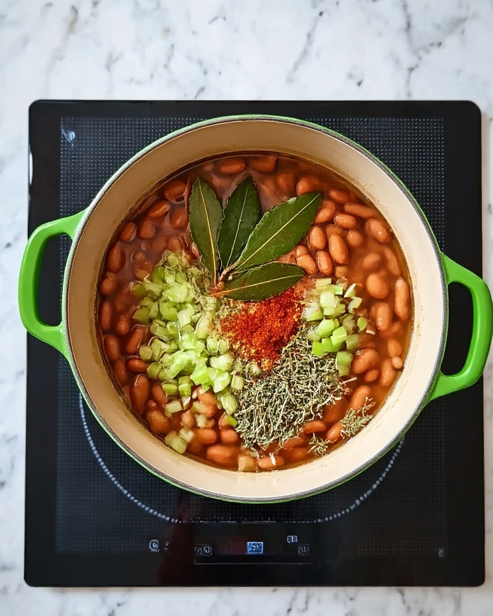 A pot with green handles is filled with reddish-brown beans sitting in water, topped with small green celery pieces, dried herbs, two dark green bay leaves, and a bit of red seasoning powder, all floating on top. The pot is on a black induction cooktop with a shiny surface, set on a white marbled countertop. photo taken with an iphone --ar 4:5 --v 7