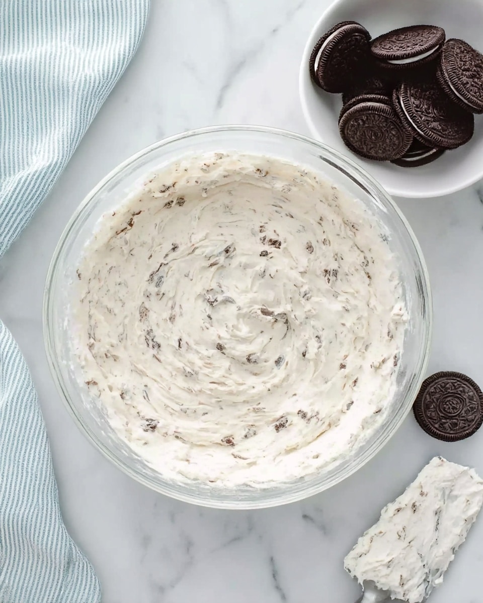 The image shows a clear glass bowl filled with a creamy white mixture that has small brown bits spread evenly throughout. The mixture has a soft, fluffy texture with swirls and peaks visible on the surface. To the right, there is a white bowl holding several whole chocolate sandwich cookies. At the bottom, part of a spatula covered with the same creamy mixture rests on a white marble surface, and a woman's hand is just out of frame. A soft blue and white striped cloth is placed on the top left side of the image. The background is a white marbled texture. photo taken with an iphone --ar 4:5 --v 7