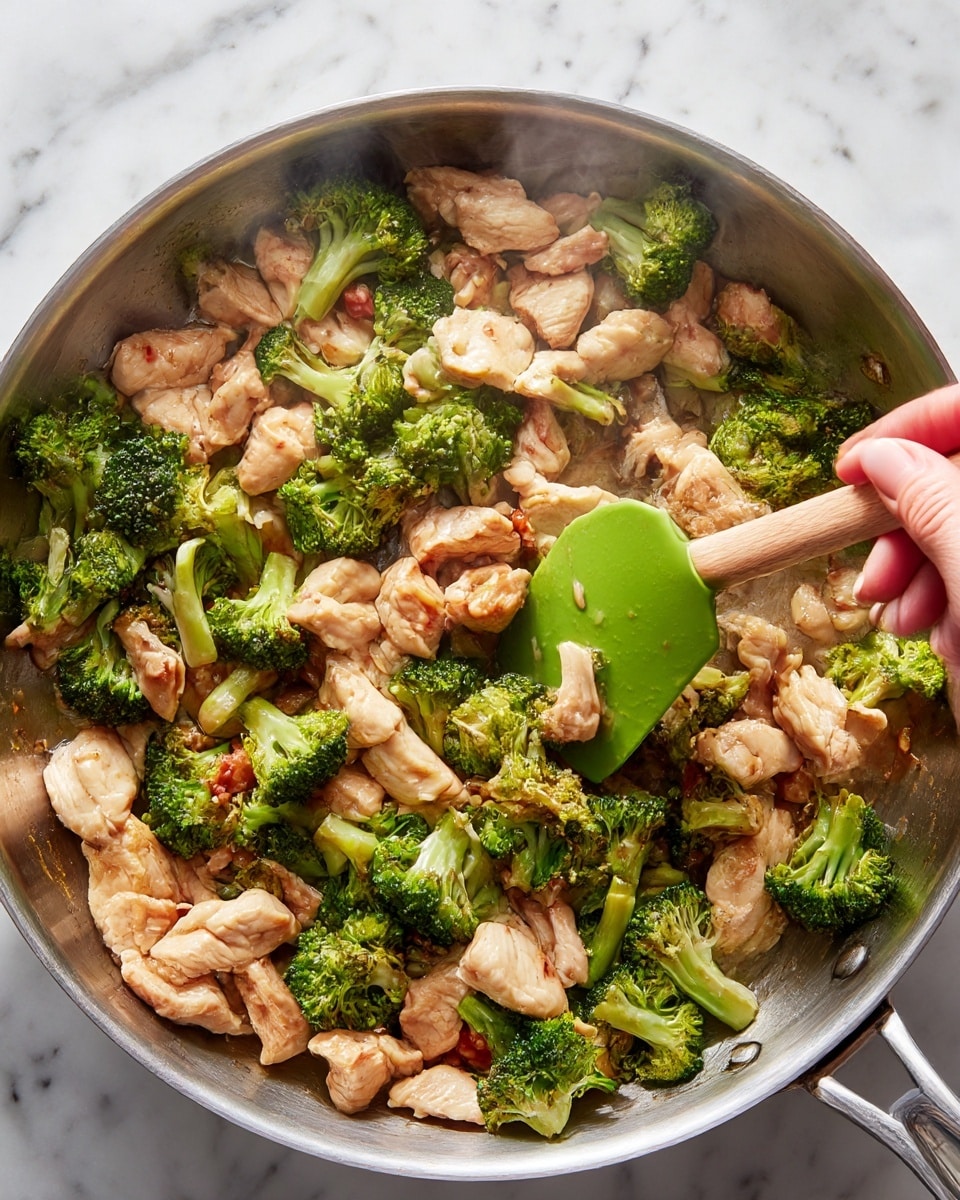 A close-up of a large silver pan filled with cooked pieces of light brown chicken and bright green broccoli florets mixed together. The chicken has a slightly browned texture, and the broccoli looks tender with some parts lightly cooked. A woman's hand holds a green spatula with a wood handle, stirring the chicken and broccoli. The pan sits on a white marbled surface, and a light steam rises from the food, showing it is hot. The photo taken with an iphone --ar 4:5 --v 7