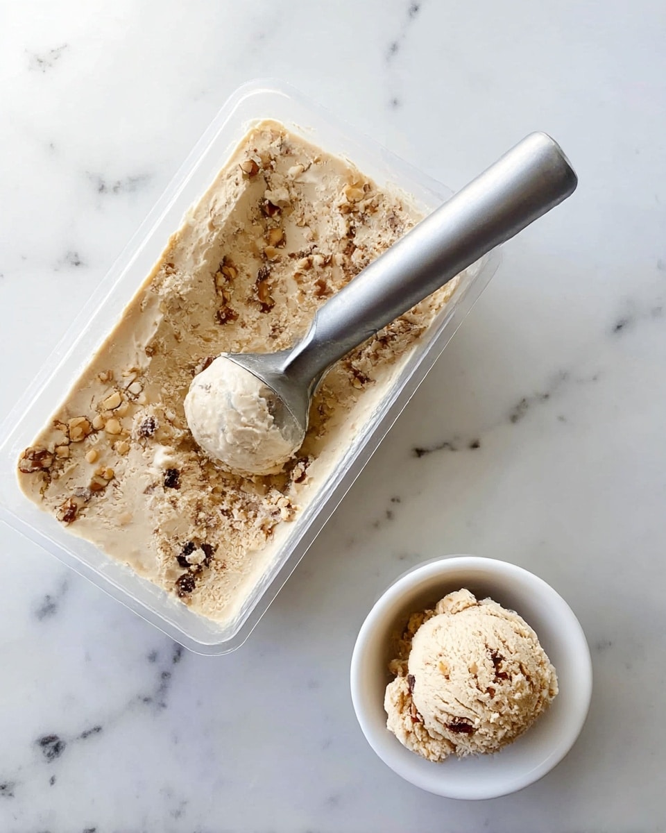 A scoop of light beige ice cream with small dark chocolate bits in it sits in a white bowl at the bottom right of the image on a white marbled surface. At the top left, a rectangular clear plastic container shows the same beige ice cream inside, with the top covered in a layer of chopped nuts and dark chocolate pieces. A shiny silver ice cream scoop with a white handle rests inside the container, partially scooping out the ice cream on the left side. The texture of the ice cream is smooth and creamy, with some rough edges where scooped. Photo taken with an iphone --ar 4:5 --v 7