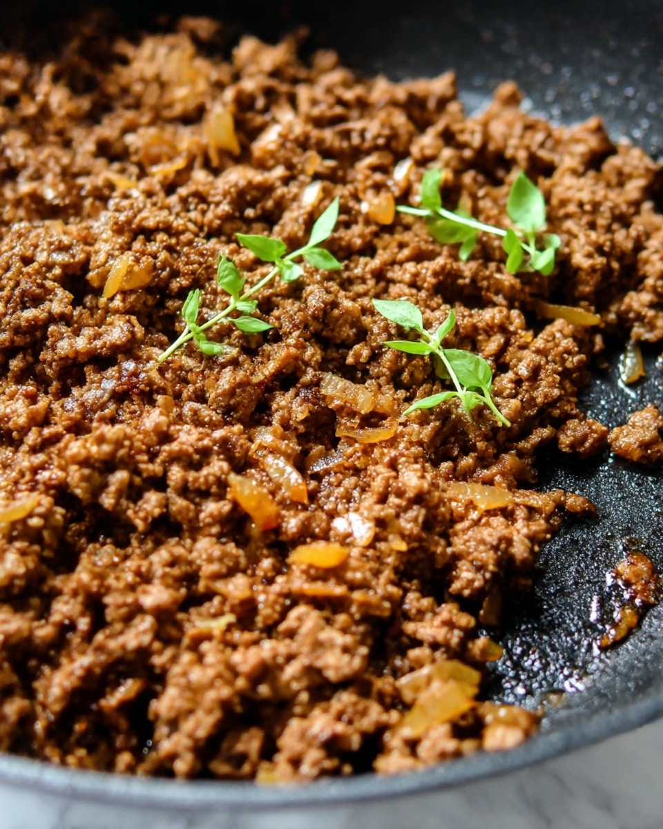 The image shows a close-up view of cooked minced meat in a black pan. The meat is browned and crumbly with small bits of caramelized onions mixed in, adding a golden color here and there. Two small sprigs of fresh green herbs lie on top of the meat, providing a contrast with the dark cooked meat. The pan surface has a slight shiny texture from the oil or juices. The background is changed to a white marbled texture. photo taken with an iphone --ar 4:5 --v 7
