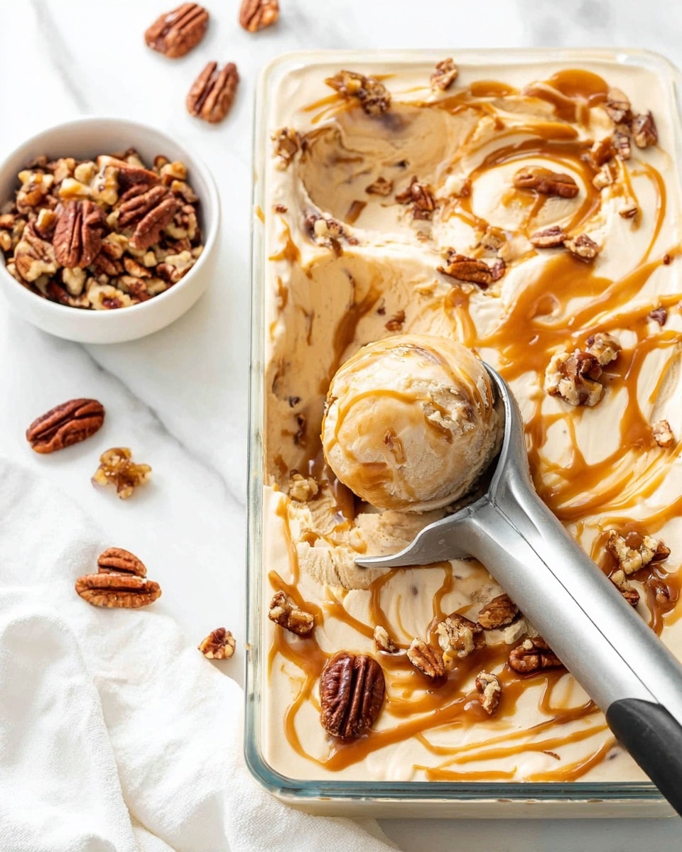 The image shows a rectangular glass pan lined with plastic wrap filled with creamy, light beige ice cream that has swirls of caramel sauce mixed through, creating flowing golden brown ribbons on the surface. There are small chunks of pecans mixed into the ice cream and sprinkled on top. A metal ice cream scoop with a black handle is lifting a rounded scoop of the ice cream from the pan. To the left, a small white bowl holds a pile of chopped pecans with several whole pecans scattered loosely around it. The background is a white marbled surface with a white cloth visible in the lower left corner. photo taken with an iphone --ar 4:5 --v 7