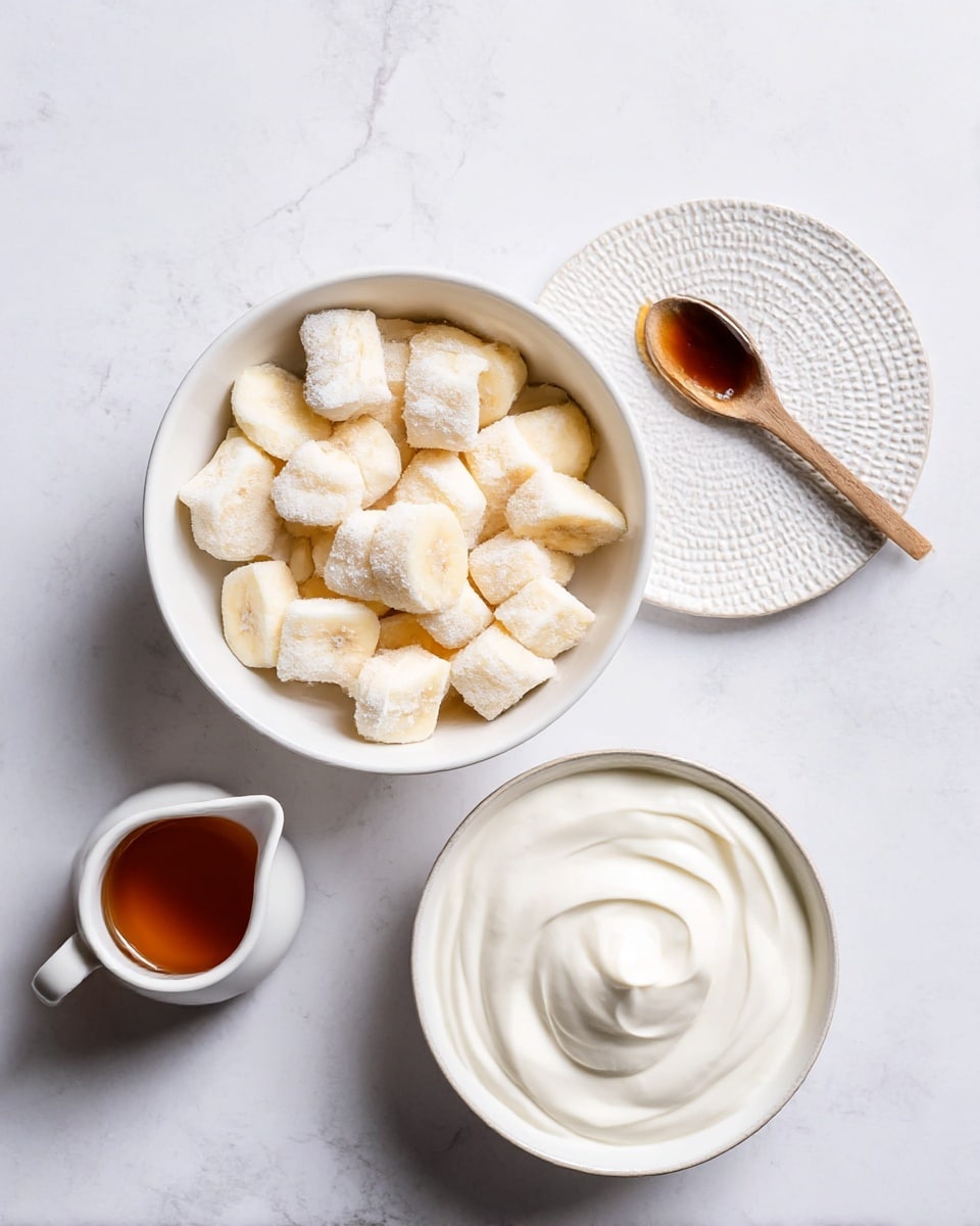 The image shows a white bowl filled with chopped frozen banana pieces that are pale yellow and frosted with ice crystals, positioned in the center on a white marbled surface. To the right is a small white plate with a textured pattern holding a wooden spoon that has a dark brown syrup. Below, there is a larger white bowl containing smooth, thick white yogurt with gentle swirls on top. At the bottom left, there is a small white pitcher filled with amber-colored syrup. All items are placed on a bright white marbled surface. Photo taken with an iphone --ar 4:5 --v 7