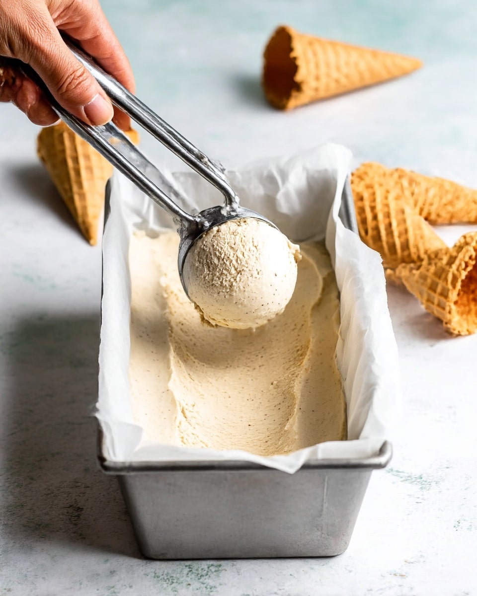 A close-up view of a thick, creamy beige ice cream in a silver metal loaf pan lined with white parchment paper. A woman's hand holds a metal ice cream scoop, lifting a round scoop of the ice cream from the pan. In the background, there are two light tan waffle cones lying on a surface with a white marbled texture. The overall scene shows the smooth texture of the ice cream with some small specks visible inside. photo taken with an iphone --ar 4:5 --v 7