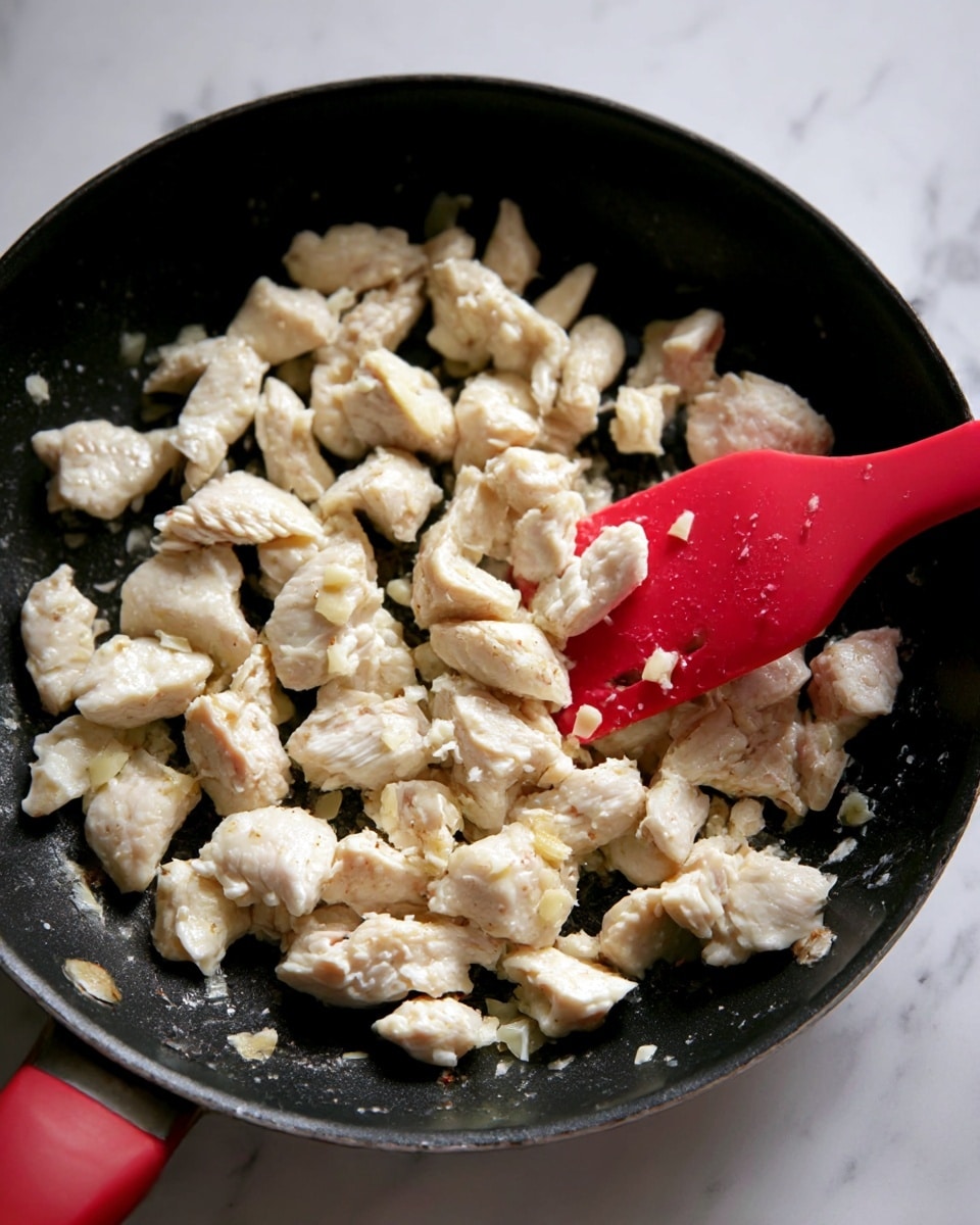 The image shows a close-up of a black frying pan with many uneven chunks of white, partially cooked chicken scattered inside. Some pieces have slight golden spots and small bits of garlic on them. A red silicone spatula is seen pushing and mixing the chicken pieces from the right side. The background surface is a white marbled texture. photo taken with an iphone --ar 4:5 --v 7