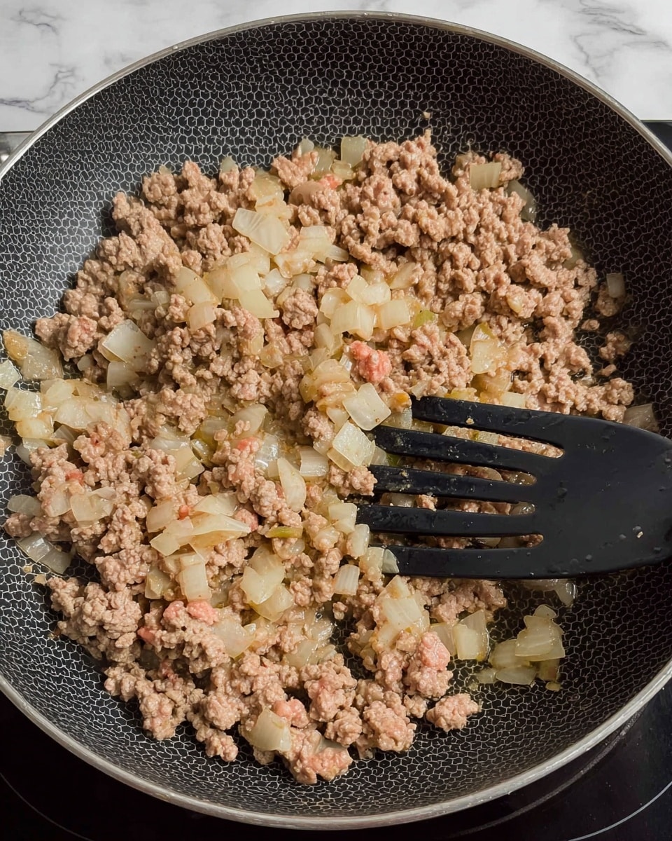 A close-up view of a black frying pan with a honeycomb pattern showing a mix of cooked ground meat and diced onions. The meat is mostly light brown with some pink spots, while the onions are soft and translucent, scattered evenly across the pan. A black slotted spatula is stirring the mixture, lifting some pieces. The pan sits on a stovetop, and the scene is set against a white marbled texture background. photo taken with an iphone --ar 4:5 --v 7