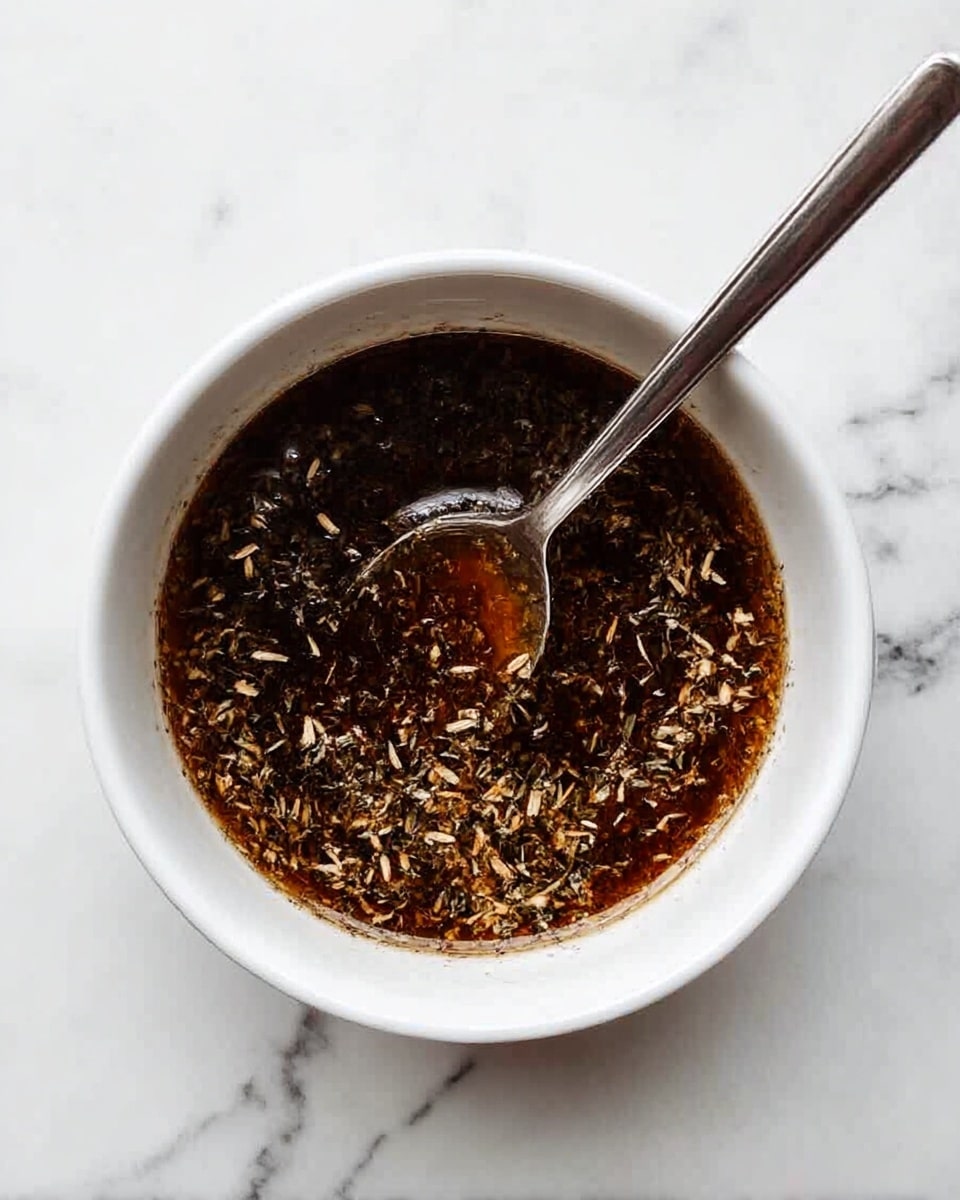 A white bowl filled with a dark brown sauce that has small light brown and beige herb flakes floating on the surface, giving it a textured look. A silver spoon is placed inside the bowl, resting on the side with its handle extending out of the bowl. The bowl sits on a white marbled surface with subtle dark gray veins. photo taken with an iphone --ar 4:5 --v 7