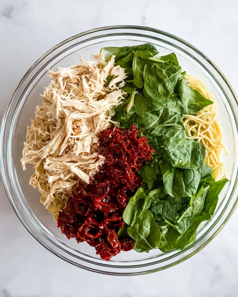 This image shows a clear glass bowl placed on a white marbled surface, filled with four main layers of fresh ingredients without mixing. The first layer is light beige shredded chicken, placed on the left side and bottom left part of the bowl with a slightly fibrous texture. Next to it, on the bottom right side, is a pile of thin, pale yellow pasta strands. On the top right side, there is a heap of bright green fresh spinach leaves with a smooth texture. At the center of the bowl, bridging between the spinach and chicken, is a cluster of dark red sun-dried tomatoes that look slightly wrinkled and shiny. The ingredients are neatly separated and vibrant, ready to be mixed. Photo taken with an iphone --ar 4:5 --v 7