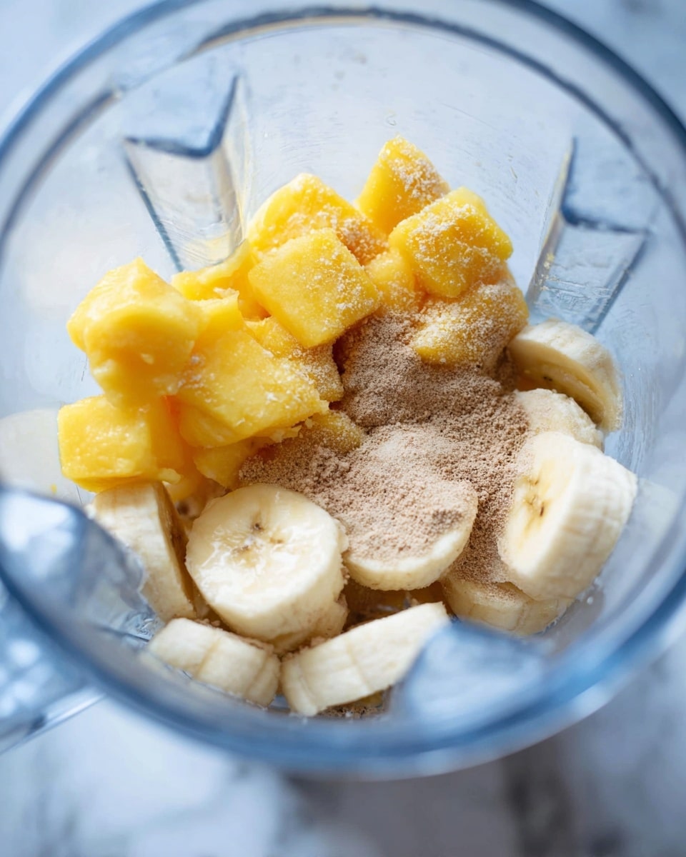 Inside a clear blender, there are three layers: at the bottom, round light yellow frozen banana slices with a slightly soft texture; above the bananas, bright yellow frozen mango chunks with a frosty coating; and on top, a light brown powder sprinkled over the fruit pieces, filling the space unevenly. The scene is set against a white marbled background. photo taken with an iphone --ar 4:5 --v 7