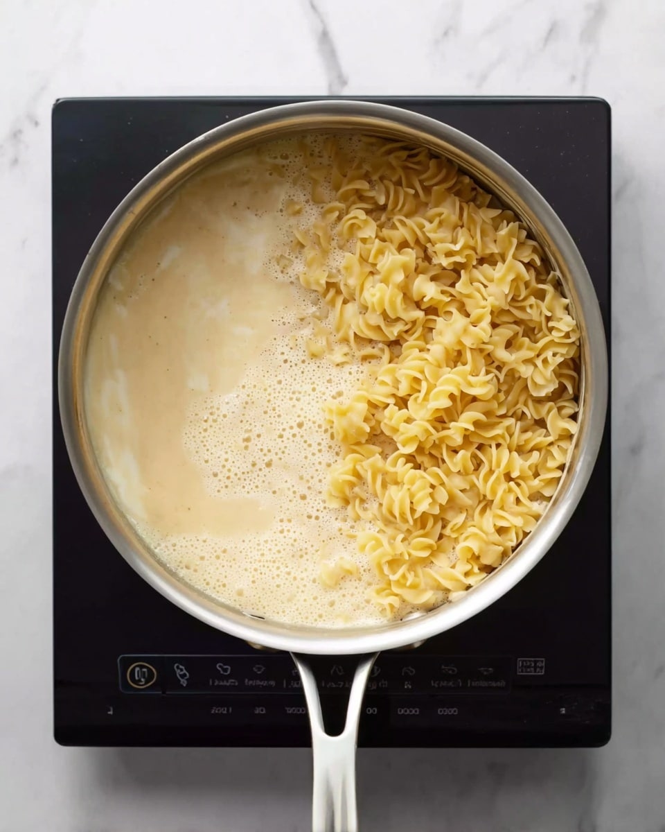 A large silver pot with two handles is shown from above, sitting on a black electric stove with buttons visible below. Inside the pot, creamy sauce fills about half of the space with a frothy texture, while on one side, half-cooked pale yellow curly pasta rests, separated from the sauce. The background is a white marbled surface. photo taken with an iphone --ar 4:5 --v 7
