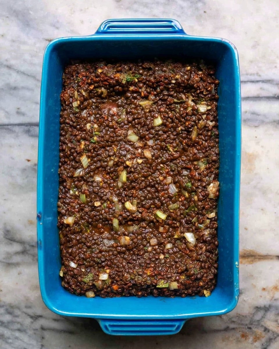 A rectangular blue baking dish filled with a cooked dark brown lentil mixture that looks slightly chunky and moist, showing small bits of lighter colored vegetables like onions mixed in. The surface of the lentils is uneven, with some visible pieces of green herbs or vegetables. The dish is placed on a white marbled surface. photo taken with an iphone --ar 4:5 --v 7
