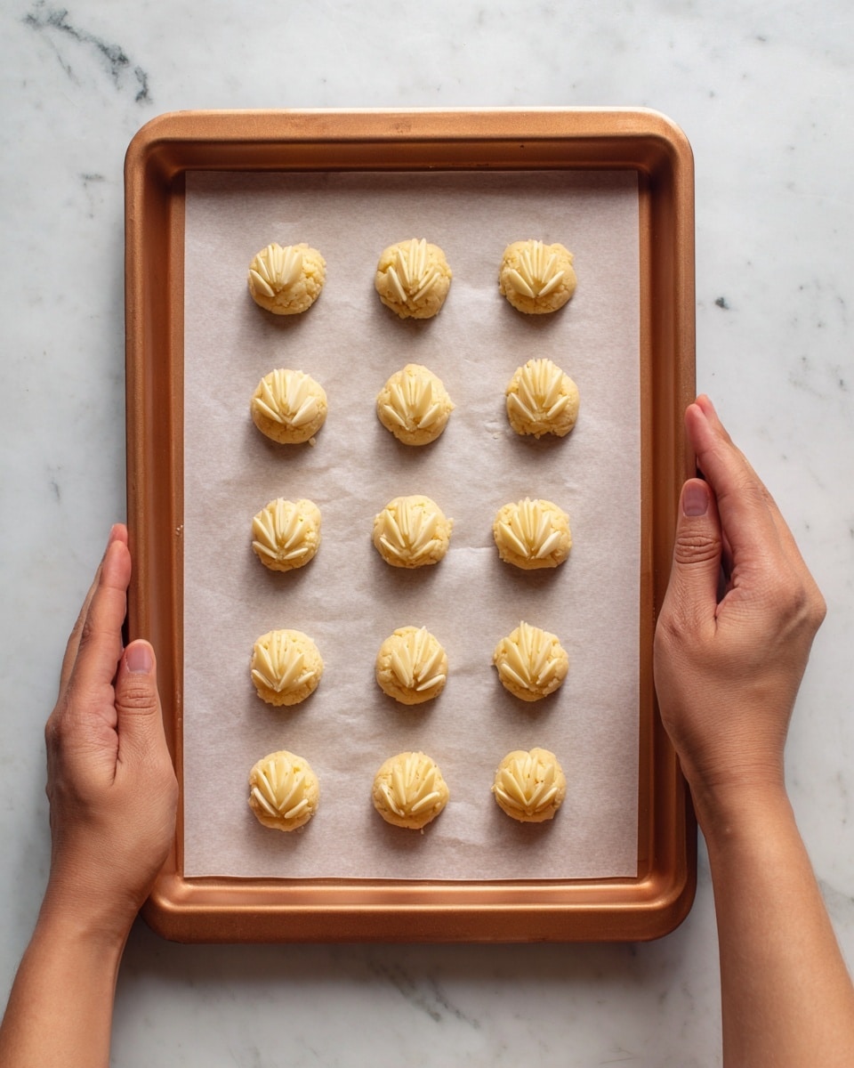 A copper baking tray holds a sheet of white parchment paper with twelve small round dough balls placed evenly in a 3x4 grid. Each dough ball is pale yellow with a rough texture, topped with three almond slices arranged in a fan shape. The tray is held at the bottom corners by two woman's hands. The whole scene is set on a white marbled surface. photo taken with an iphone --ar 4:5 --v 7