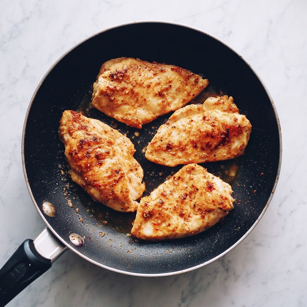 A black frying pan holds four pieces of cooked chicken, each piece showing golden-brown spices and a light crust. The chicken pieces are spread evenly, and the surface of the pan has small drops of shiny oil. The background surface is a white marbled texture. photo taken with an iphone --ar 4:5 --v 7