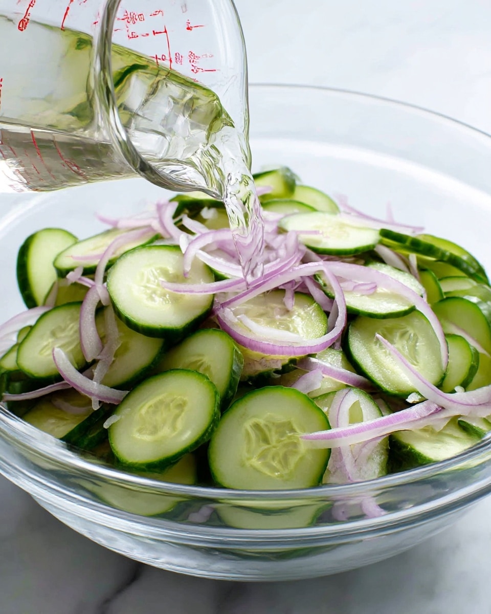 A clear glass bowl on a white marbled surface filled with thinly sliced cucumber rounds showing a light green inside and darker green edges, layered with thin, soft purple onion slices spread evenly on top and inside. A clear liquid is being poured from a clear measuring cup with red markings into the bowl, catching light reflections. The overall look is fresh and crisp, with a mix of smooth cucumber surfaces and slightly translucent onion strips. photo taken with an iphone --ar 4:5 --v 7