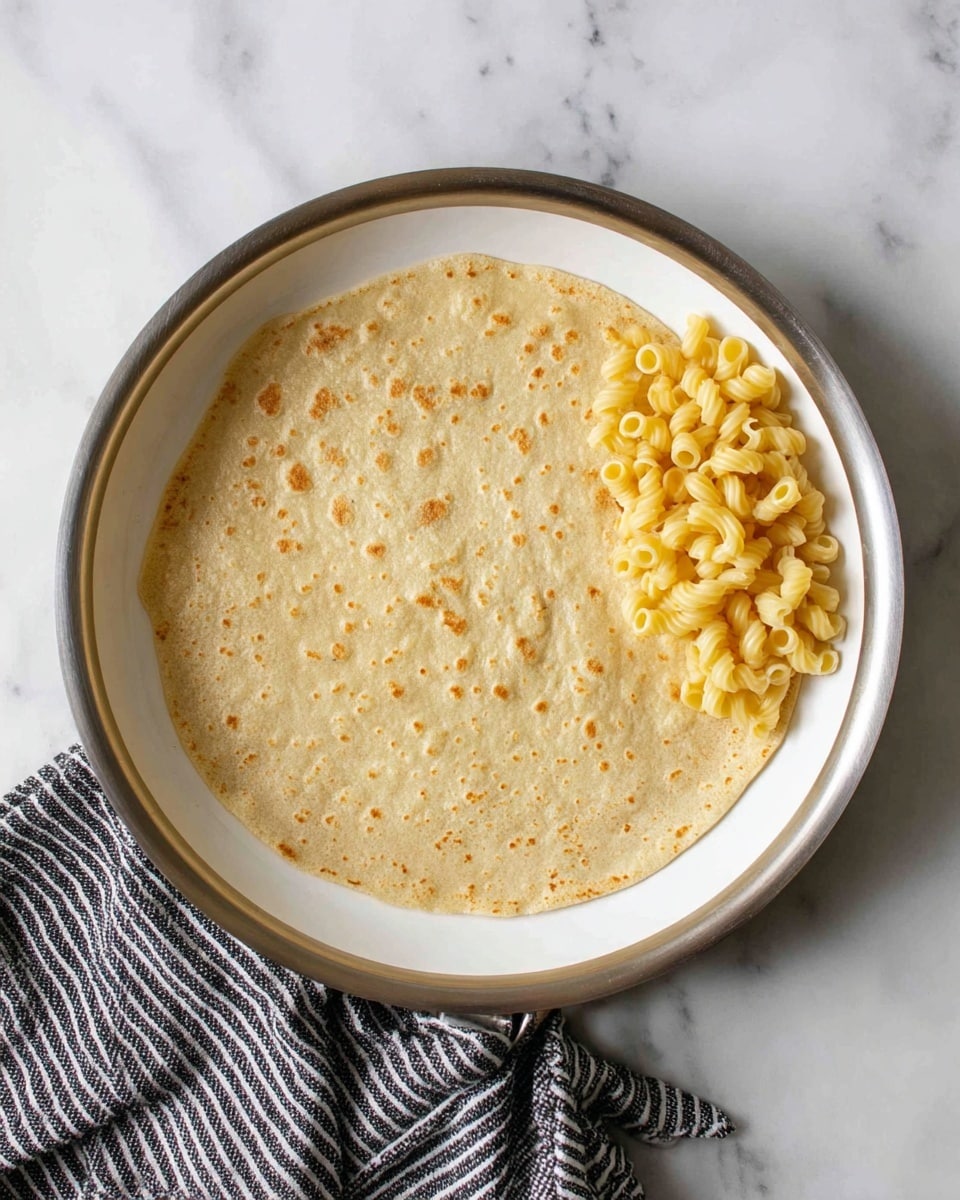 A white pan holds a flat light brown tortilla with a slightly uneven surface and small darker specks, covering most of the pan’s base. On the right side of the tortilla, there is a small pile of yellow macaroni pasta with a soft texture and a few curved elbow shapes. The pan rests on a white marbled surface and a black-and-white striped cloth is partially visible on the lower right side. photo taken with an iphone --ar 4:5 --v 7