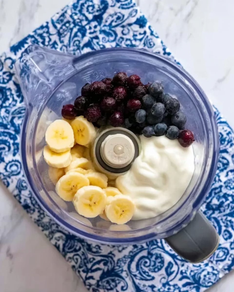 The image shows a clear blender jug placed on a blue and white patterned cloth with a white marbled background. Inside the blender, there are several colorful layers: on the left side, sliced yellow bananas; on the top right, small dark purple berries; and on the bottom right, a thick white creamy layer, likely yogurt, all arranged separately but touching each other. The blender's blade is visible in the center, surrounded by the ingredients. Photo taken with an iphone --ar 4:5 --v 7