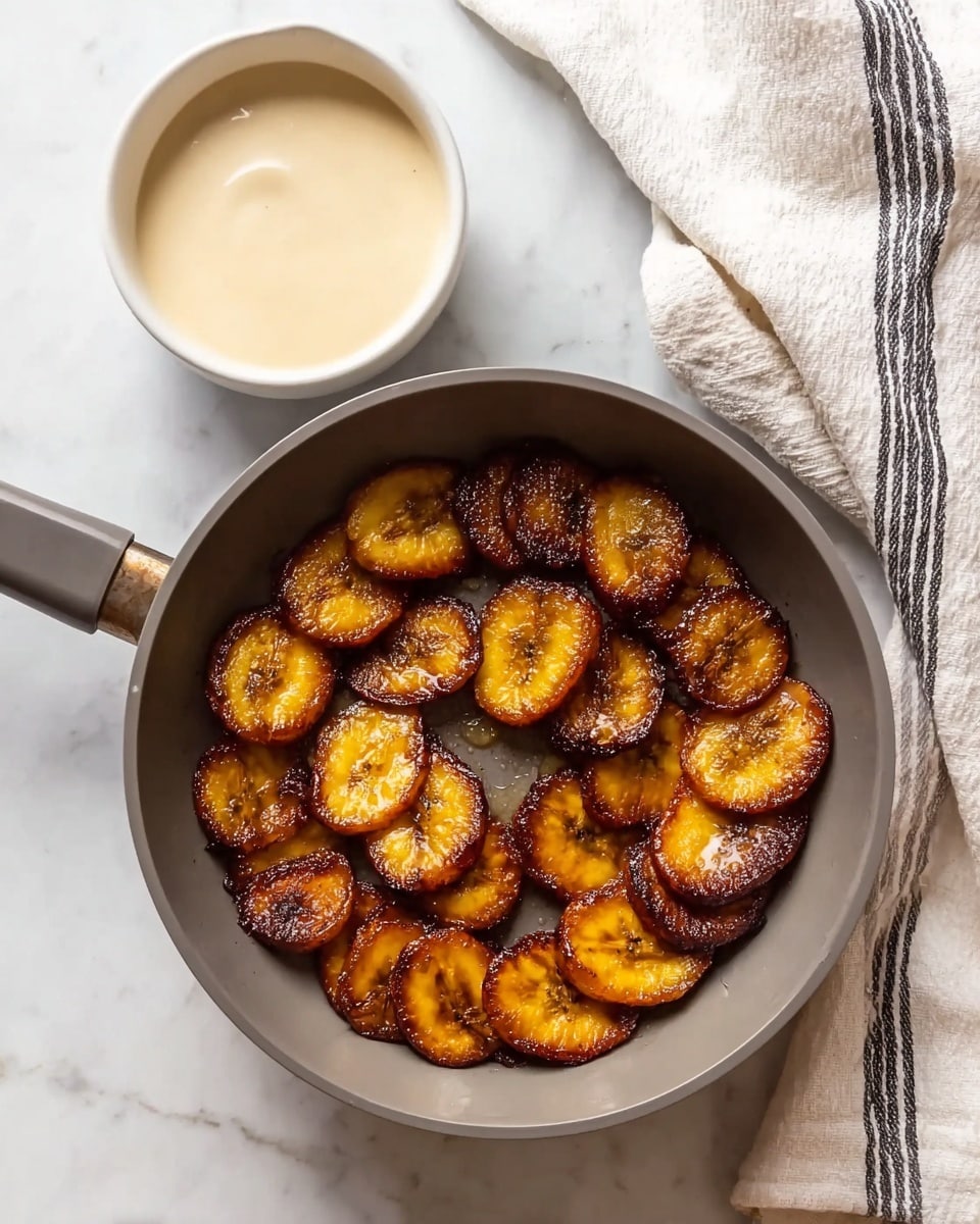 A round gray pan contains one layer of golden brown fried plantain slices arranged in a neat circle, each slice showing caramelized edges and a soft, shiny texture. To the top left of the pan sits a single white bowl filled with cream-colored smooth sauce. The pan and bowl rest on a surface with a white marbled texture, and to the right of the pan is a white cloth with black stripes, loosely folded. The lighting is soft, making the colors of the food warm and rich. photo taken with an iphone --ar 4:5 --v 7