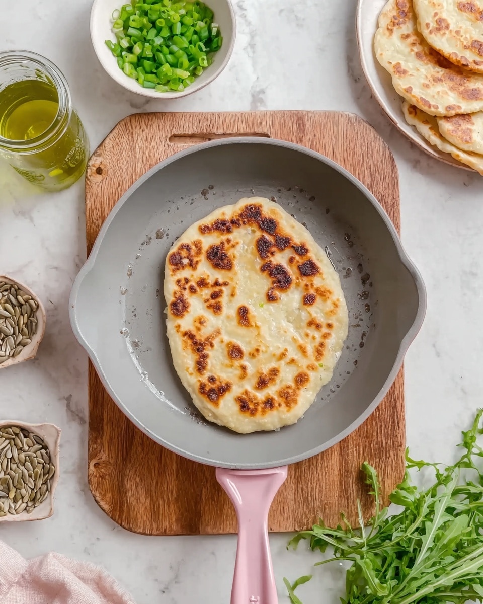 A single flatbread with a golden brown, unevenly toasted surface lies in the center of a pink-handled grey pan, which sits on a wooden cutting board. Around the pan, there is a small white bowl filled with chopped green onions on the upper left, a glass jar containing green oil to its left, a white dish with sunflower seeds, and a small bunch of fresh green herbs and arugula on the lower right. Part of a white plate with more flatbreads is visible in the top right corner, all arranged on a white marbled surface. photo taken with an iphone --ar 4:5 --v 7