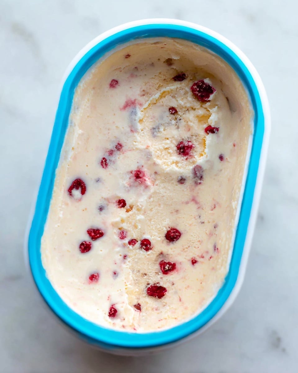 The image shows a light beige ice cream with visible scattered small red berries mixed inside. The ice cream fills a white container with a bright blue rim at the top edge. The surface of the ice cream is smooth with a few scooped-out spots revealing a creamy texture and more berries embedded inside. The container is placed on a white marbled surface. Photo taken with an iphone --ar 4:5 --v 7