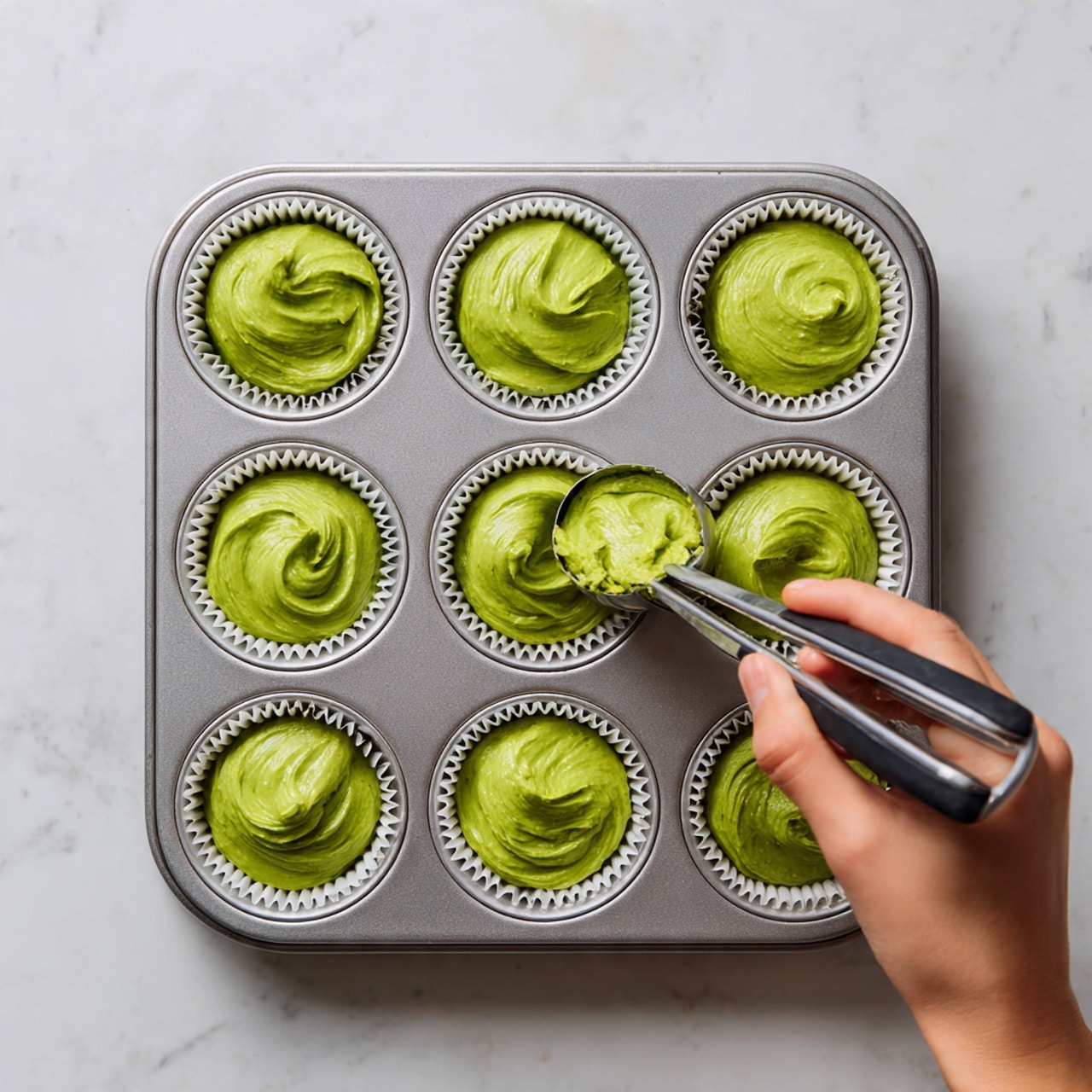 The image shows a metal muffin tray with six white paper liners filled with bright green batter. Each liner holds a soft, swirled mound of smooth, thick batter with a slightly glossy texture. The tray is placed on a white marbled surface. A woman's hand is holding a silver scoop with black handles, adding more green batter into one of the liners. The batter looks fresh and creamy, filling each liner evenly but with some peaks from the scooping motion. Photo taken with an iphone --ar 4:5 --v 7