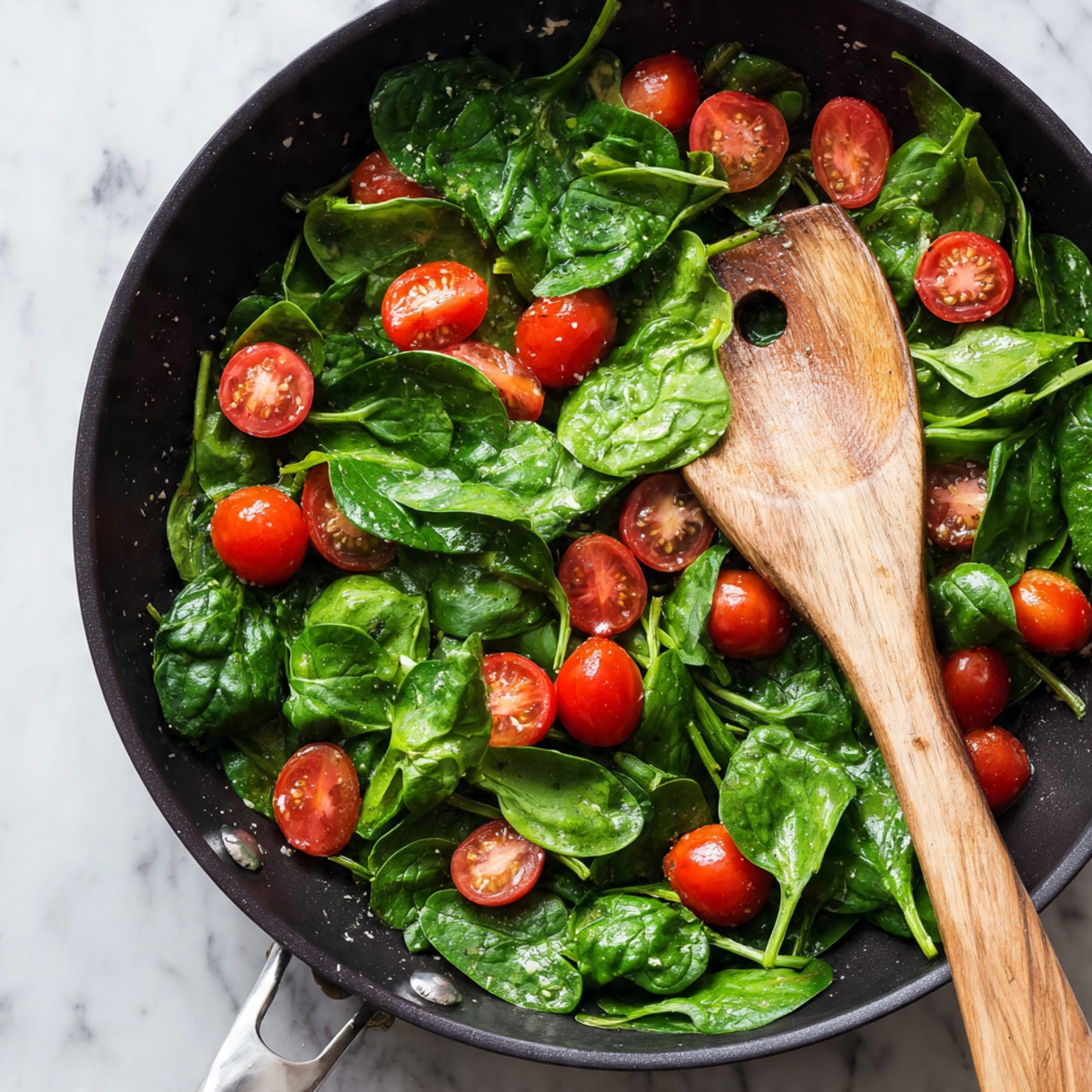 The image shows a black pan filled with fresh green spinach leaves scattered across it, with bright red cherry tomatoes cut in half and whole ones mixed throughout. A light wooden spoon with a hole in the middle lies on top of the spinach, angled diagonally from the right side. The spinach leaves have a fresh, slightly wet texture, and the tomatoes are shiny and smooth. The pan edges are white, placed on a white marbled surface photo taken with an iphone --ar 4:5 --v 7