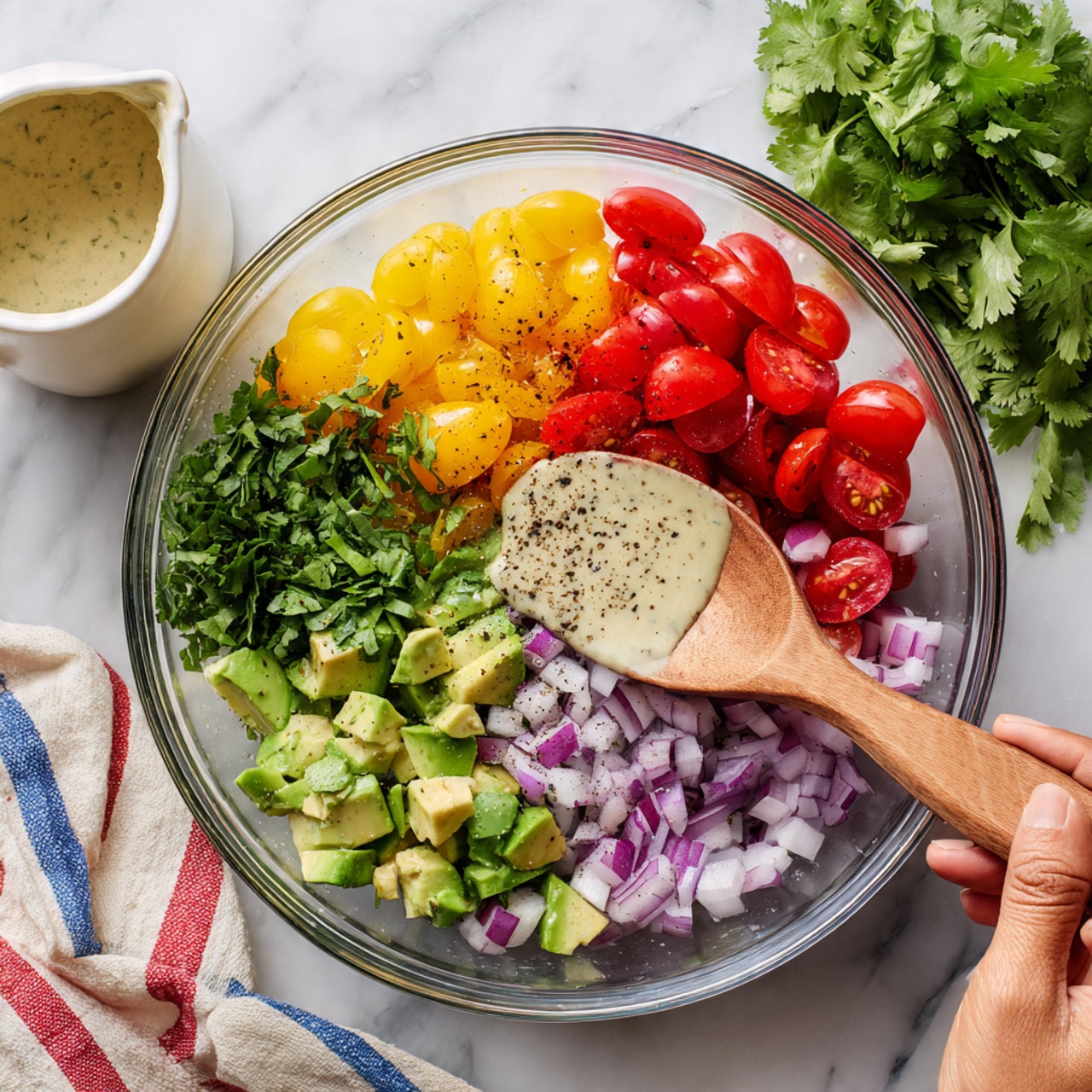 A clear glass bowl contains several layers of fresh ingredients on a white marbled surface. The bottom layer is made of mixed halved cherry tomatoes in red, yellow, and purple colors. Above them are chopped green peppers and diced purple onions. On one side, there are small pieces of green avocado with a creamy texture. Fresh green herbs like cilantro fill the empty space in the bowl. A woman's hand holds a wooden spoon on the right side, mixing the contents, while another woman's hand pours a light beige creamy dressing with black pepper from a white small pitcher on the left side. Fresh cilantro leaves lie in the background alongside a folded striped cloth in red, white, and blue colors photo taken with an iphone --ar 4:5 --v 7