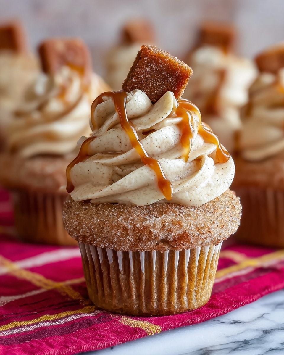 A single cupcake is shown standing on a red cloth with yellow stripes over a white marbled surface. The cupcake has three main layers: the bottom layer is the soft brown cake with a sugar-coated outer edge that adds a crystalline texture. The middle layer is a generous swirl of creamy, pale beige frosting with little specks of spice visible throughout. The top layer is a small, golden-brown square piece coated in sugar, placed at an angle. Thin ribbons of caramel sauce are drizzled over the frosting and the sugar-coated square, adding a shiny contrast. In the blurred background, more cupcakes with the same details appear on the white marbled surface. photo taken with an iphone --ar 4:5 --v 7